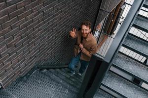Hello gesture. Man in khaki colored jacket and in jeans is on the stairs on the building outdoors photo