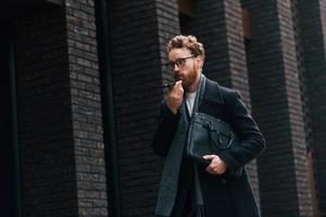 Walking near building. Stylish man with beard and in glasses is outdoors photo