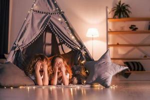 Covering eyes by hands and laying down. Two little girls is in the tent in domestic room together photo