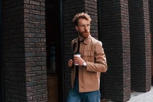 Coffee break. Stylish man with beard in khaki colored jacket and in jeans is outdoors near building photo