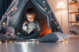 Using flashlight to read the book. Cute little girl playing in the tent that is in the domestic room photo