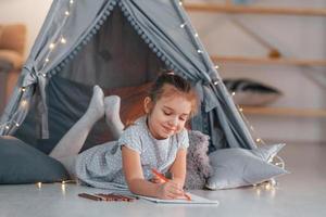 With writing equipment. Cute little girl playing in the tent that is in the domestic room