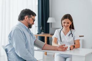 Female doctor is measuring blood pressure of the man. Professional medical worker in white coat is in the office photo