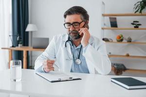 Man giving information by phone. Professional medical worker in white coat is in the office photo