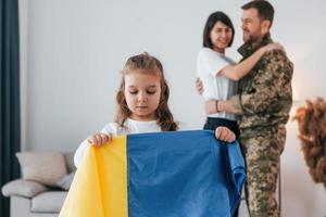 Girl standing in front of her parent and holding Ukraine flag. Soldier in uniform is at home with his wife and daughter photo