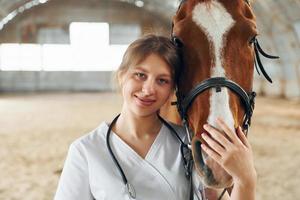 Portrait of female doctor that is in white coat is with horse on a stable