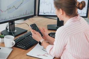 Using smartphone. Female stock broker in casual clothes is working in the office by pc photo