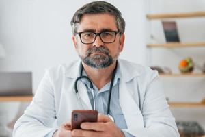 Male doctor is sitting with phone in hands. Professional medical worker in white coat is in the office photo