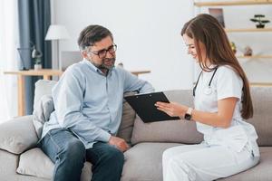 Female doctor is sitting with mature customer on the sofa. Professional medical worker in white coat is in the office photo