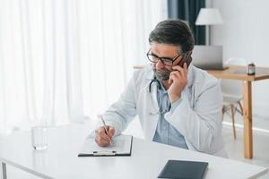 hombre dando información por teléfono. trabajador médico profesional con bata blanca está en la oficina foto