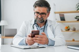 Male doctor is sitting with phone in hands. Professional medical worker in white coat is in the office photo