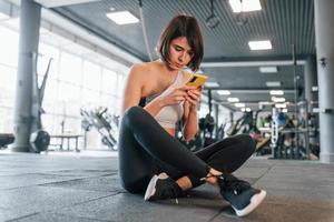Using smartphone. Woman in sportive clothes with slim body type is in the gym photo
