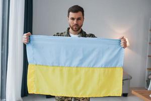 Soldier in uniform holding Ukrainian flag in hands at home photo