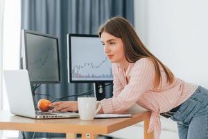 de pie con manzana en la mano y mirando la computadora portátil. una corredora de bolsa con ropa informal está trabajando en la oficina por pc foto