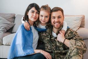 la bandera ucraniana está en los hombros. soldado en uniforme está en casa con su esposa e hija foto