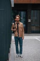 vista frontal. hombre elegante con barba en chaqueta de color caqui y en jeans está al aire libre cerca del edificio foto