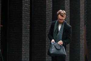Black colored wear and bag. Stylish man with beard and in glasses is outdoors near building photo