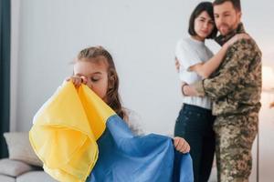 Girl standing in front of her parent and holding Ukraine flag. Soldier in uniform is at home with his wife and daughter photo