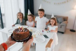 Woman holding cake with candles. Celebrating birthday. Group of children is together at home at daytime photo