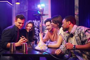 Talking with each other when sitting by the table with hookah. Group of friends having fun in the night club together photo