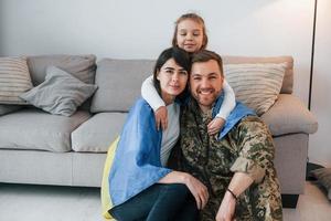 Ukrainian flag is on the shoulders. Soldier in uniform is at home with his wife and daughter photo