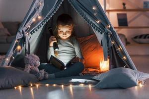 Using flashlight to read the book. Cute little girl playing in the tent that is in the domestic room photo