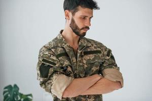 brazos cruzados. soldado en uniforme está de pie en el interior contra la pared blanca foto