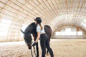 At daytime. A young woman in jockey clothes is preparing for a ride with a horse on a stable photo