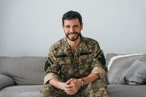 Soldier in uniform is indoors in the domestic room. Sitting on the sofa photo