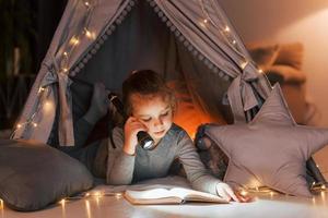 Reading book at evening. Cute little girl playing in the tent that is in the domestic room photo