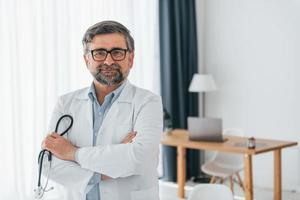 de pie y sosteniendo estetoscopio. trabajador médico profesional con bata blanca está en la oficina foto