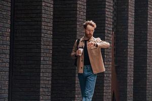 Coffee break. Stylish man with beard in khaki colored jacket and in jeans is outdoors near building photo