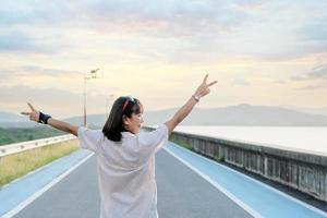 Back view of a female tourist rising hands up in the air. Woman traveler standing on street of dam with a sunset background on a vacation trip. Asian lady enjoying nature on holiday. Freedom concept photo