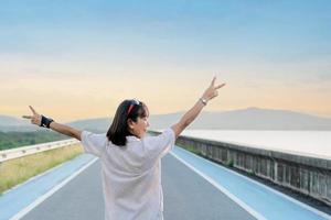 Back view of a female tourist rising hands up in the air. Woman traveler standing on street of dam with a sunset background on a vacation trip. Asian lady enjoying nature on holiday. Freedom concept