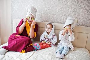 madre con hijas en un albornoz con una toalla en la cabeza cuida la piel en casa, pepinos en los ojos. foto