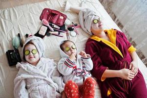 Mother with daughters in a bathrobe with a towel on her head take care of the skin at home, cucumbers on eyes. photo