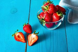 Fresh red strawberries in transparent bowl and small kitchen towel in blue wooden background photo