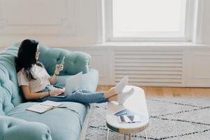 Sideways shot of relaxed female student uses modern technologies while doing homework, uses web sources for making project, sits on comfortable sofa against cozy interior, leans legs on desktop photo