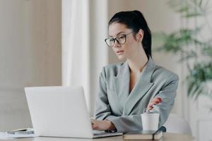 Serious female entrepreneur in eyeglasses, has dark hair combed in pony tail, works on business project, concentrated at laptop computer wears grey formal costume uses information from web resources photo