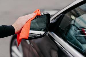 Close up of mans hand wiping water on black car with microfiber cloth. Focus on auto side mirror. Transporation self service photo