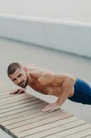 Vertical shot of muscular bearded man stands in plank pose trains chest and arms muscles has hard workout for keeping athletic body shape poses outdoor. Active lifestyle and motivation concept photo