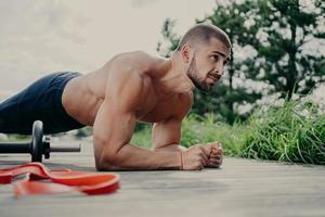 Photo of motivated bodybuilder does plank exercise, stands on elbows, uses sport equipment for workout, concentrated into distance, has muscular body, enjoys training outdoor. Morning fitness concept