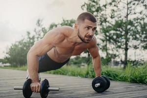 Muscular male bodybuilder does push up with barbells stands in palnk pose with naked body, trains determination, goes in for sport regularly. Sport, fintess, powerlifting and bodybuilding concept photo