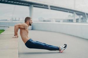 Sideways shot of muscular man does reverse push up exercise, trains arms, keeps perfect body and good physical shape, has active workout outdoor, wears sport trousers and sneakers, poses on bridge photo