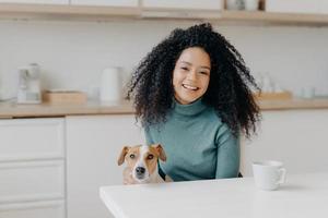 Pretty young lady with Afro hairstyle plays with dog, wears comfortable jumper, drinks coffee or tea in kitchen, going to have walk together, rejoice good weather outdoor. People, animals, spare time photo