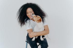linda mujer feliz con el pelo rizado recibe un beso de jack russell terrier siente amor por su mascota favorita se complace en compañía de un perro sentado en una silla contra un fondo blanco. amor entre dueño y animal. foto