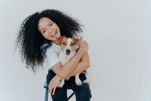 Pleasant looking curly girl tilts head, smiles happily, embraces favourite dog, has good time with pet, wears casual t shirt and jeans, sits on comfortable chair against white background. Relationship photo