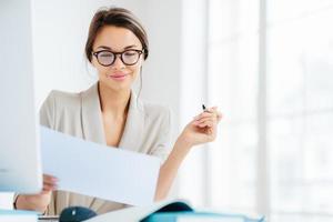 Concentrated successful businesswoman looks attentively in paper, studies terms of contract, holds pen, writes in documentations, dressed formally, poses at desktop against white spacious interior photo