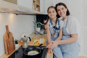 encantadora madre e hija con delantales se abrazan y sonríen alegremente, fríen huevos en una estufa moderna en la cocina, usan una sartén, preparan un delicioso desayuno. concepto de familia, niños, maternidad y cocina. foto