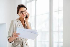 Horizontal shot of business lady discusses details of contract, works in office, concentrated on information from accountings, holds paper documents during telephone conversation, dressed formally photo
